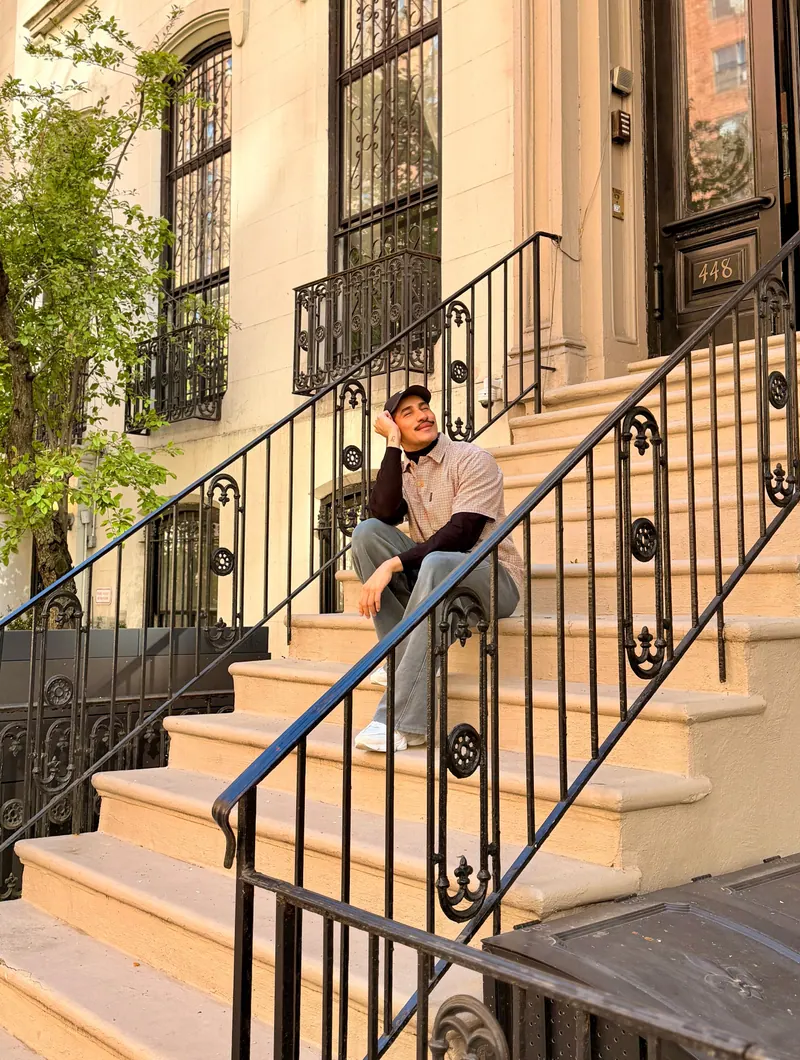 Ives sentado na escadaria de um prédio clássico de Nova York, sorrindo