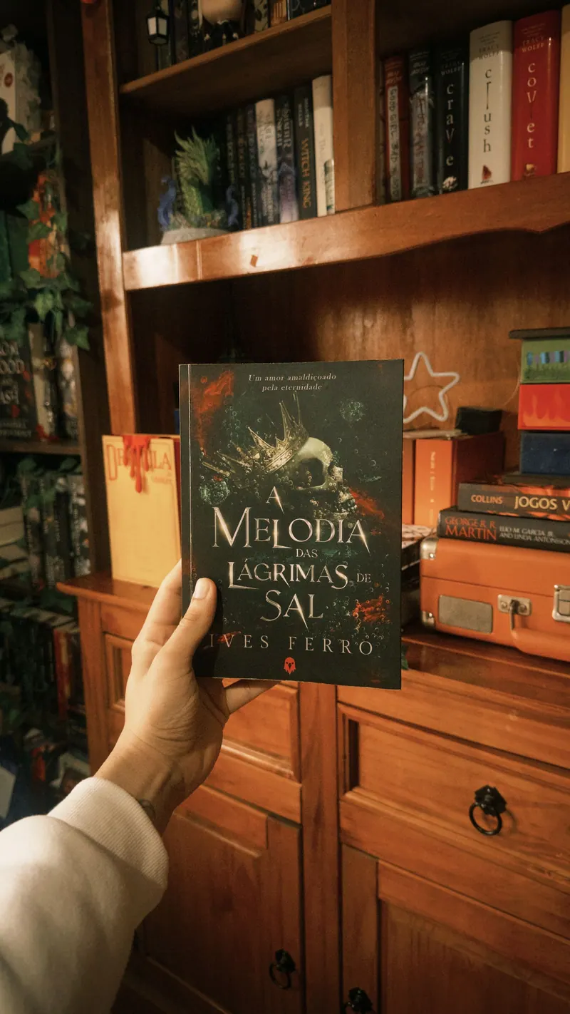 Mão segurando o livro A Melodia das Lágrimas de Sal, de Ives Ferro, em frente a uma estante