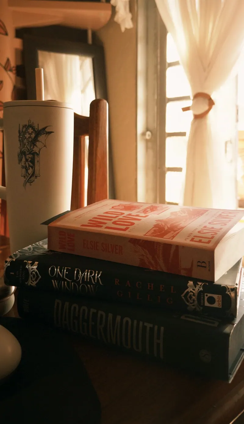 Pilha de livros perto de uma janela com cortina clara e uma caneca